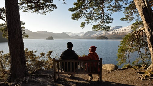 A couple sit on a bench looking out towards Derwent Water and the snow capped hills on a clear winters day